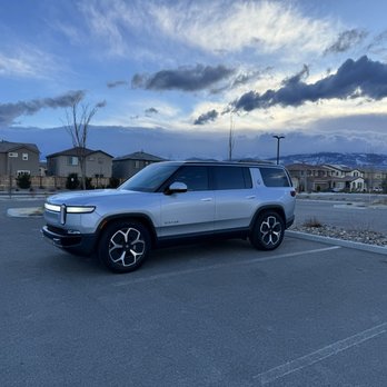 Premier Window Tinting - 20% ceramic tint up front and 15% in rear - A silver Rivian R1S SUV is parked in an empty parking lot with marked spaces. The background features residential houses, leafless trees, and mountains under a partly cloudy sky during dusk. The scene is illuminated by natural light, with a calm and quiet atmosphere. - Home Window Tinting Near Me - Reno, Nevada