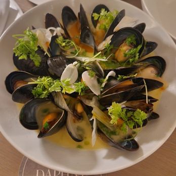 Daphne - A white bowl contains steamed mussels with open shells, topped with chopped garlic and fresh herbs such as cilantro. The dish is presented on a wooden table with a Daphne restaurant coaster visible underneath. In the background, there is a partially visible salad plate with greens and other ingredients. - Seafood Near Me - Toronto, Ontario