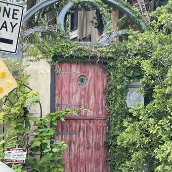 Explore Tours and Pick Ups - A red wooden door with metal hinges and a small circular window is set in a garden with dense green foliage and vines. - Historical Tours Near Me - St. Augustine, Florida