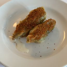 Squash Blossom - Two squash blossom appetizers, coated in a crispy, seasoned breading, are presented on a white ceramic plate. The dish appears to be part of a seafood-focused menu at a waterfront dining establishment, with a small amount of sauce or melted butter underneath. The background includes a glass of water and a white tablecloth, indicating a formal dining setting.