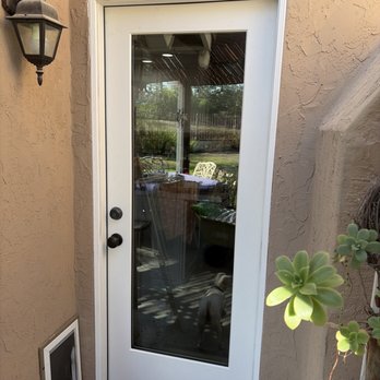 The Unhinged Door - Rear Door - A white exterior door with a large glass panel is mounted on a stucco wall, with a black handle and lock, and a small pet visible through the glass. - Door Sales/Installation Near Me