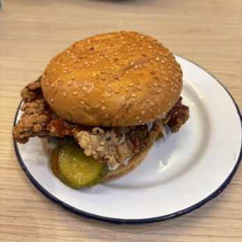 Chimac - Classic chicken burger - A chicken burger features a fried chicken fillet, sliced pickles, and sauce inside a sesame seed bun. The burger is served on a white plate with a black rim, placed on a light wooden table. In the background, there is a bowl of food, possibly rice or another dish, on the table. - Chicken Wings Near Me - Dublin 2, D