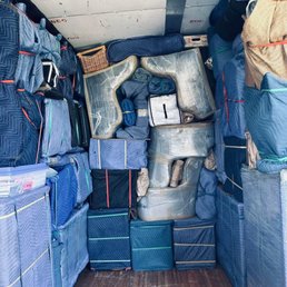 Multiple stacks of blue and gray moving blankets, padding, and packing materials are arranged inside a truck. The blankets are secured with straps, and some are wrapped in plastic. A small wicker basket and a white box are also visible among the packing supplies. The truck has a wooden floor and a metal wall with some markings and scratches.