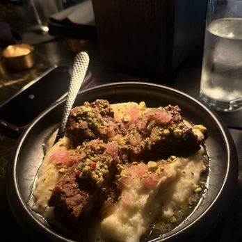 Dearly Beloved - Short rib - A plate features short rib meat on a bed of mashed potatoes, garnished with crushed pistachios, in a dark restaurant setting. - Cocktail Bars Near Me - Chicago, Illinois