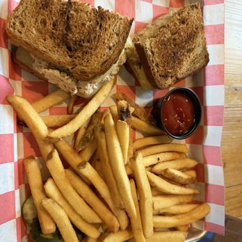 Eli's - Tuna melt - A meal served on a metal tray lined with red and white checkered paper includes a tuna melt sandwich with toasted bread, French fries, sliced pickles, and a small cup of ketchup. The sandwich appears to contain tuna salad and melted cheese, with the fries being golden and crispy. The pickles are sliced and placed at the corner of the tray, and a serrated knife is visible at the top of the image. - Breakfast & Brunch Near Me - Natick, Massachusetts