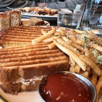 Valentina's - A close-up of a restaurant meal featuring a grilled cheese sandwich with toasted bread, crispy fries topped with herbs, and a small cup of ketchup. In the background, there are additional plates with pizza slices and bread, along with water glasses and a person wearing a gray jacket sitting at the table. The setting appears to be a casual dining restaurant. - Italian Near Me - Dublin, Ohio