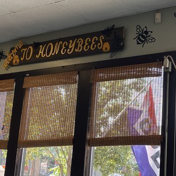 Honeybees - A sign with the text "Welcome to Honeybees" is mounted above a window with bamboo blinds, featuring bee and flower decorations. - New American Near Me - Geneva, Ohio