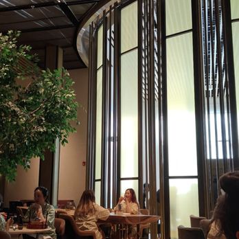 Go Place - Dining area - The image shows the interior of a restaurant with high ceilings and large, curved windows allowing natural light to fill the space. A sizable indoor tree is positioned on the left side, adding greenery to the setting. Several groups of people are seated at tables, engaged in conversation or dining. The decor includes modern architectural elements, with a mix of wooden and metal features, creating a contemporary atmosphere. - Day Spas Near Me - Markham, Ontario