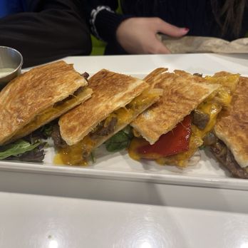 Fusion Crunch - Malaysian Quesadilla - A rectangular white plate holds four quesadillas cut into halves, revealing melted cheese, beef, and vegetables inside. The quesadillas are golden brown and crispy, garnished with lettuce leaves on the side. A small metal container of sauce is placed at the corner of the plate. The background shows a table with partially visible people and a casual dining setting. - Burgers Near Me - Bloomington, Illinois