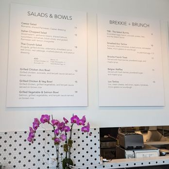 Cafe Select - The image shows a cafe interior with two large menu boards mounted on a light-colored wall. The left board lists salads and bowls, including Caesar Salad, Italian Chopped Salad, Thai Crunch Salad, and various grilled chicken and salmon bowls, with prices ranging from $11 to $19. The right board features brunch options such as TSB - The Select Burrito, Smashed Avo Tartine, Brioche French Toast, Belgian Waffles, and Lox Tartine, with prices from $9 to $13. Below the menu, a potted orchid with pink flowers is placed on a black countertop, which also holds a container with utensils and some electronic devices. The background includes a partial view of the kitchen area. - Coffee & Tea Near Me - Fort Lee, New Jersey