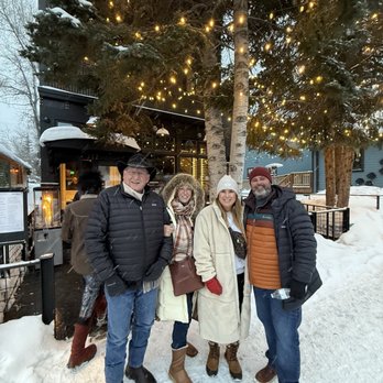 Two Twelve - Front view - Four people dressed in winter coats, hats, and boots stand on snow-covered ground outdoors. They are positioned in front of a large tree decorated with string lights, with a building visible behind them. The scene suggests a winter outing or gathering in a cold climate. - New American Near Me - Crested Butte, Colorado