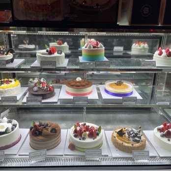 Paris Baguette - Cakes - A multi-tiered glass display case contains a variety of decorated cakes, each with different toppings such as strawberries, berries, chocolate, and fruit garnishes. The cakes are arranged on white trays with labels, indicating flavors and types. The display is well-lit, highlighting the colorful and detailed decorations of each cake, and is situated in a bakery or pastry shop setting. - Coffee & Tea Near Me - Hackensack, New Jersey
