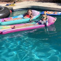 Two young girls are relaxing on inflatable loungers in a backyard pool, wearing swimsuits and sunglasses, with pool toys and a fence in the background.
