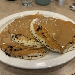 Addictive delight pancakes - Three pancakes with chocolate chips are arranged on a white oval plate. They are topped with a layer of peanut butter and sprinkled with powdered sugar. The pancakes are placed on a beige table, with a glass of water and some utensils visible in the background.