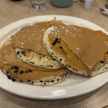 Maxfield's Pancake House - Addictive delight pancakes - Three pancakes with chocolate chips are arranged on a white oval plate. They are topped with a layer of peanut butter and sprinkled with powdered sugar. The pancakes are placed on a beige table, with a glass of water and some utensils visible in the background. - Breakfast & Brunch Near Me - Milwaukee, Wisconsin