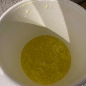 New Dragon - Egg drop Egg Flower Soup - A white disposable cup contains a yellowish liquid with shredded ingredients floating inside, likely egg drop soup. The cup is placed on a white surface, and part of a foil wrapper is visible at the bottom right corner of the image. The background includes a textured wall and a shadow cast across the cup. - Chinese Near Me - Albuquerque, New Mexico