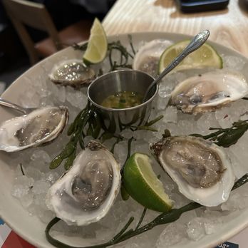 Mariscos San Pedro - A round white plate contains several raw oysters on the half shell, placed on a bed of crushed ice. The oysters are garnished with lime wedges and seaweed, with a small metal container of dipping sauce in the middle, accompanied by a spoon. The setting appears to be a restaurant, with a wooden table and people in the background. - Mexican Near Me - Chicago, Illinois