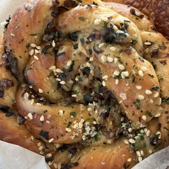 Third Cliff Bakery - Mushroom chive bun - A baked pastry with a golden-brown, flaky crust is shown, topped with chopped herbs, sesame seeds, and small dark bits, likely mushrooms or olives. The pastry appears to be seasoned and garnished, indicating a savory flavor profile. It is placed on parchment paper, suggesting it is freshly baked and ready for serving, from Third Cliff Bakery in Boston, MA. - Cafes Near Me - Boston, Massachusetts