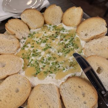 Capri Restaurant - Whipped ricotta with honey - A round plate contains whipped ricotta cheese drizzled with honey and sprinkled with chopped pistachios. The plate is surrounded by toasted bread slices arranged in a circular pattern. The setting appears to be in a restaurant, with a dark wooden table and a stack of small white plates and napkins nearby. - Pizza Near Me - Loves Park, Illinois