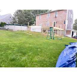 A backyard with a grassy lawn features a green ladder and construction materials, including wooden framing and a blue tarp. The background shows a brick house with multiple windows and neighboring houses, suggesting ongoing outdoor construction or renovation work.