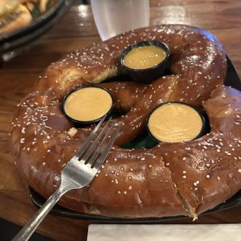 More Brewing - The huge pretzel. I put the fork in there to show the size. - A large pretzel with a golden-brown crust and coarse salt is placed on a black plate. Three small containers of cheese sauce are arranged on top of the pretzel. A fork is partially inserted into the pretzel. In the background, there is a glass of water and a blurred dish, with a wooden table surface visible. - Brewpubs Near Me - Villa Park, Illinois