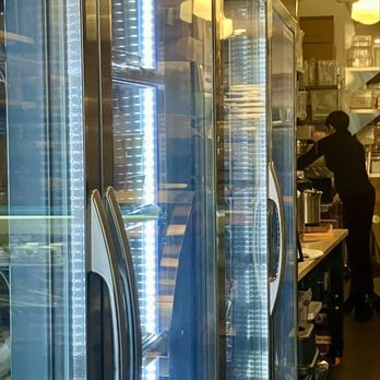 Electric Beets - A tall, glass-fronted refrigerator with metal handles and illuminated interior LED lighting is shown. The refrigerator is located in a commercial kitchen or deli setting, with a person working in the background. The environment includes shelves, containers, and kitchen equipment, indicating a food service area. - Vegan Near Me - Brooklyn, New York