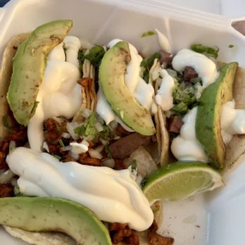 La Costa - 4 chicken tacos with cream & avo. - Three tacos filled with seasoned meat, topped with avocado slices, sour cream, chopped onions, and cilantro, served in a white takeout container. Lime wedges are included for added flavor, and the presentation emphasizes fresh ingredients and typical taco toppings. - Mexican Near Me - Tracy, California