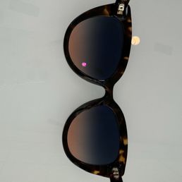 Hinge fixed - A pair of tortoiseshell sunglasses with dark tinted lenses is positioned on a light, smooth surface. The frames have a rounded shape with a pattern typical of tortoiseshell material. The image captures the sunglasses from a top-down perspective, highlighting the design details and reflections on the lenses. The background is plain, emphasizing the sunglasses as the main subject.