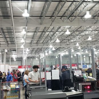 Costco - The image shows the interior of a Costco wholesale store with numerous customers shopping and queuing at checkout counters. The store has high ceilings with exposed metal beams and bright lighting. Shelves stocked with products line the background, and customers are seen pushing shopping carts and carrying items. The checkout area includes cash registers and staff assisting customers, with a clear view of the store's spacious layout. - Tires Near Me - Pflugerville, Texas