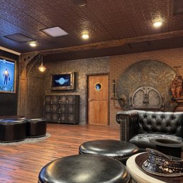 A themed escape room interior featuring leather seating, brick and metal walls, and industrial-style decor with a large circular door.