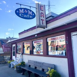 Street View / Entrance - parking is limited out front of the cafe - The image shows the front of M & M Cafe with a neon sign, windows displaying menu signs, and outdoor seating with benches and plants.