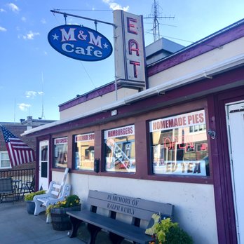M & M Cafe - Street View / Entrance - parking is limited out front of the cafe - The image shows the front of M & M Cafe with a neon sign, windows displaying menu signs, and outdoor seating with benches and plants. - Cafes Near Me - Monticello, Wisconsin
