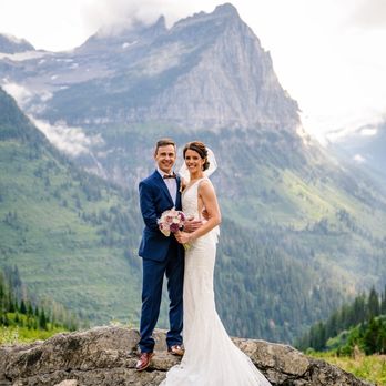 Bella Tailor - A bride and groom stand on a large rock formation outdoors, with a mountain range and forested landscape in the background. The groom wears a blue suit with a bow tie, and the bride wears a white lace wedding gown holding a bouquet of flowers. The scene captures a wedding moment in a natural, mountainous setting. - Sewing & Alterations Near Me - Denver, Colorado