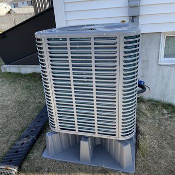 Aldo’s HVAC - ECOER Condensor 1 - The image shows a sizable outdoor HVAC unit, likely an air conditioner or heat pump, placed on a concrete pad beside a house with white vinyl siding. The unit has a protective grille and is connected to the house's electrical system. The surrounding area includes a grassy yard and a black drainage or cable conduit on the ground. The background features neighboring houses and a clear sky. - Heating & Air Conditioning/HVAC Near Me - Medford, Massachusetts