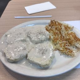 2 Biscuits and Gravy with Hashbrowns - A plate with biscuits covered in white gravy and topped with black pepper, served alongside crispy hashbrowns, on a wooden table at a casual restaurant.