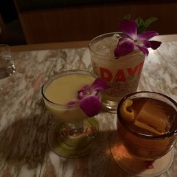 Round 2 - Three cocktails with flower garnishes are arranged on a marble table in a dimly lit environment. One drink is in a coupe glass with a purple flower, another in a tall glass with ice and a purple flower, and the third in a short glass with a lemon twist. A small glass of water is also visible in the background. The setting appears to be a bar or restaurant, with a warm, intimate atmosphere.