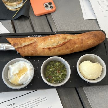 6Smith - A loaf of crusty bread is placed on a black rectangular plate, with three small metal bowls containing butter, herb sauce, and a creamy spread. The plate is on a table with menus, a glass of drink, a smartphone, and other items visible around it. - Seafood Near Me - Wayzata, Minnesota