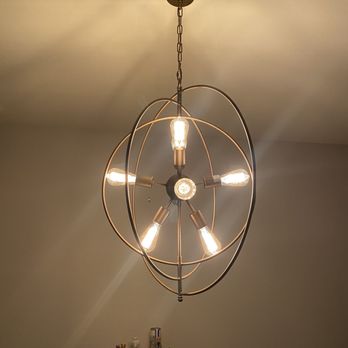 Affordable Electrical Services - Light fixture - A ceiling-mounted chandelier with a spherical metal frame design holds five exposed filament bulbs, providing illumination. The fixture is suspended by a chain from a plain ceiling, and a small bar cart with bottles and glasses is visible below. The setting appears to be an indoor space with neutral-colored walls. - Electricians Near Me