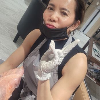 Dazzle Nails - A woman with dark hair, wearing a face mask and gloves, is seated at a table in an indoor setting that appears to be a salon or workspace. She is making a peace sign with her right hand and looking at the camera. The background includes chairs, a counter with various items, and a wall with a map or decorative mural. The environment suggests a professional or service-oriented setting. - Nail Salons Near Me - Sandy, Oregon