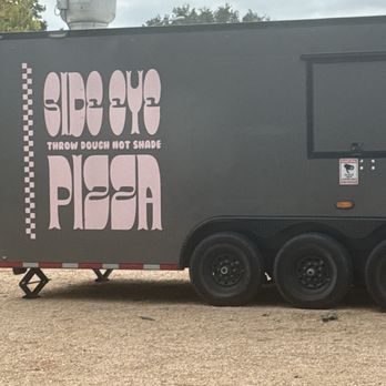 WhataKabob - A dark-colored food truck features a large sign with the text "Side Eye Pizza" and the slogan "Throw Dough Not Shade." The truck is parked on a gravel surface with trees and cloudy sky in the background. - Persian/Iranian Near Me - Austin, Texas