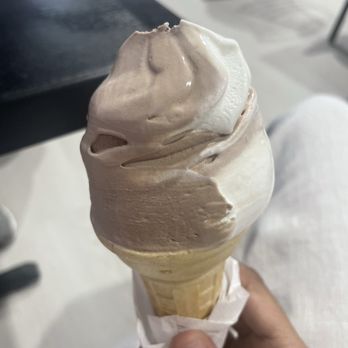 Mister Softee - Twist - A person is holding a waffle cone with two swirled soft-serve ice cream flavors, one light-colored and the other darker, possibly vanilla and chocolate or coffee. The cone is wrapped in a paper napkin, and the background shows a blurred indoor setting with tables and chairs. The focus is on the ice cream cone, which appears creamy and well-formed. - Ice Cream & Frozen Yogurt Near Me - Saint Augustine, Florida