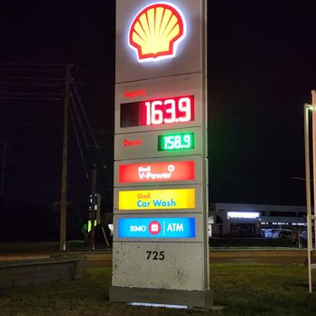 Shell - Signage - The image features a Shell gas station sign illuminated against a dark night sky. The sign displays fuel prices: 163.9 for regular and 158.9 for diesel. It also lists services including Shell V-Power, Shell Car Wash, and an ATM operated by BMO. The sign is situated on a grassy area near a road, with some background structures and streetlights visible. - Gas Stations Near Me - Burlington, Ontario
