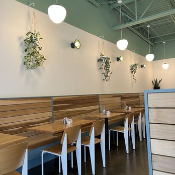 Beau's cafe - Interior - The image shows the interior of a café with a high ceiling and exposed metal beams. The wall is decorated with hanging plants and wall-mounted lights. There are round pendant lights hanging from the ceiling, providing illumination. Wooden tables with matching chairs are arranged along the wall, separated by a low partition. The overall design emphasizes simplicity and natural elements, with a clean and modern aesthetic. - Breakfast & Brunch Near Me - Lexington, Kentucky
