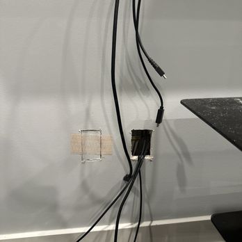 FixTman - Washington - Multiple black electrical cables hang loosely from an open wall outlet, which is embedded in a wall with a visible patch of wall repair. The cables are not connected to any devices, and the wall appears to be in the process of electrical work or repair. The setting is indoors, with a dark floor and a black countertop partially visible on the right side of the image. - Furniture Assembly Near Me