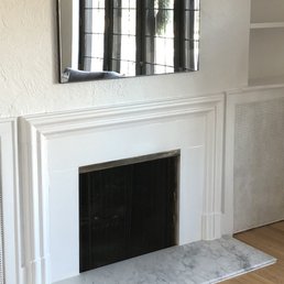 The image shows a white fireplace with a marble hearth situated against a wall. Above the fireplace, there is a mirror with rounded edges reflecting a window with bars and outdoor scenery. To the right of the fireplace, built-in shelves are visible, and the floor is hardwood with a patterned rug partially visible at the bottom of the image.