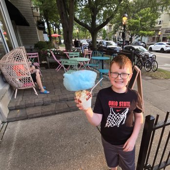 Hey Sugar - Cloud 9 - A young boy with glasses, wearing a black Ohio State shirt and shorts, stands on a sidewalk holding a large cotton candy with a blue fluffy top. Behind him, there are pastel-colored tables and chairs outside a dessert shop, with a person sitting on a pink chair. The scene is set on a tree-lined street with parked cars and bicycles, and the time appears to be late afternoon or early evening. - Coffee & Tea Near Me - GENEVA, Illinois