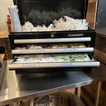 FarmHand Kchn - Cute. They use mechanic tool boxes. - A black Husky coffee urn with multiple labeled compartments is positioned on a stainless steel table. The top compartment contains plastic bags, and the front drawers are labeled with blue tape for utensils, sugar in the raw, splenda, sugar, and stevia. The setting appears to be a restaurant or cafe, with a wooden countertop and other items visible in the background. - Breakfast & Brunch Near Me - St. Augustine, Florida