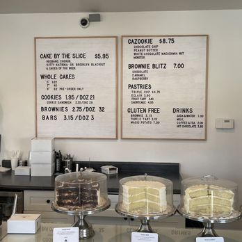 The Chocolate - Menu - The image features a dessert cafe counter with four cakes displayed under glass domes, including chocolate, vanilla, and layered options. Behind the counter, there are two menu boards listing prices for cakes, cookies, brownies, bars, and beverages. The cafe interior includes a black countertop, a cash register, and various supplies, with a doorway visible on the right side. - Desserts Near Me - Orem, Utah