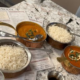 Butter Chicken - Three bowls of Indian curry, garnished with herbs and spices, are arranged on a marble table alongside two bowls of white rice. The bowls are metallic, and each has a spoon. Menus from Tikka TapRoom are partially visible underneath the bowls, indicating the restaurant's offerings. The setting suggests a dining experience focused on Indian fusion cuisine.