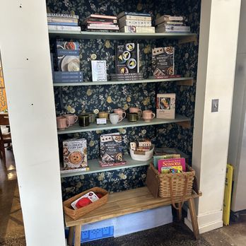 Spilt Milk - Items being sold - A shelving unit with three shelves holds various cookbooks, mugs, and small kitchen items. The background features dark floral wallpaper. A wooden bench in front of the shelves has baskets containing additional items. The setting appears to be inside a retail store or cafe. - Bakeries Near Me - Oak Park, Illinois